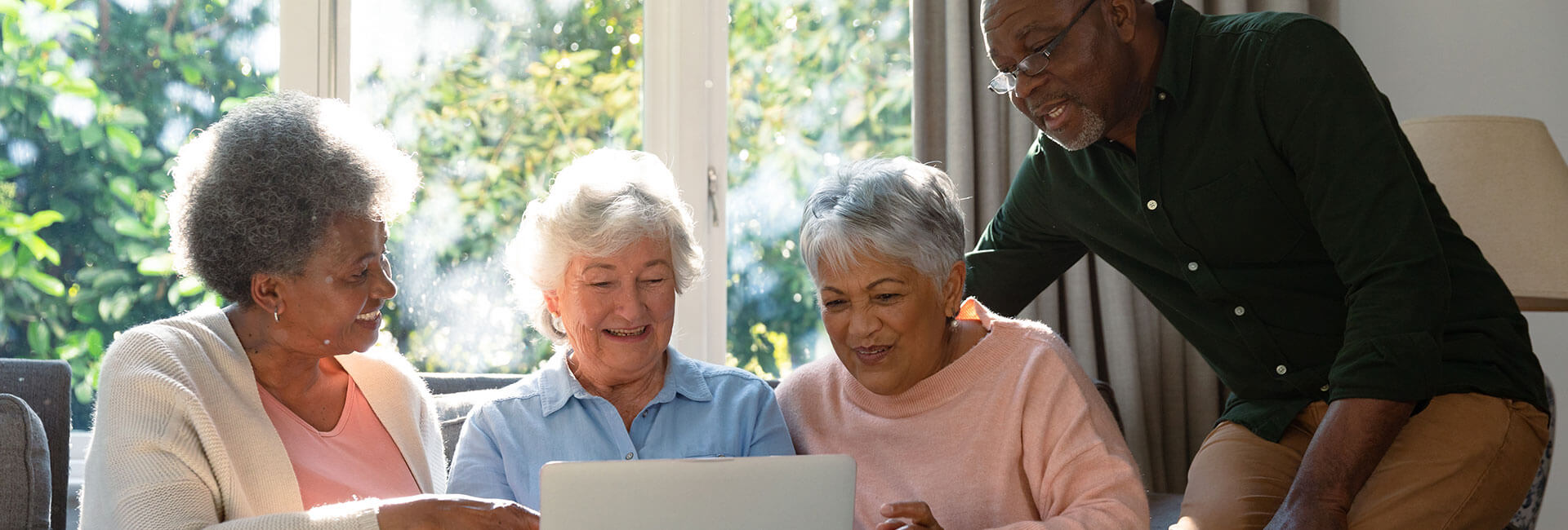 Three happy diverse senior woman and african american male friend sitting on sofa and using laptop. retirement lifestyle relaxing at home with technology.
